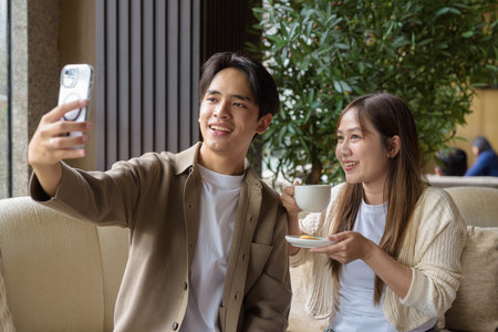 Young Asian couple drinking coffee and taking selfie with mobile phoneの写真素材