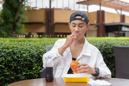 Portrait of non binary gay man eating fried chicken and drinking cola outdoors during summerの写真素材