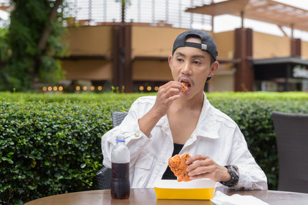 Portrait of non binary gay man eating fried chicken and drinking cola outdoors during summerの写真素材