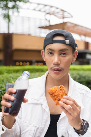 Portrait of non binary gay man eating fried chicken and drinking cola outdoors during summerの写真素材
