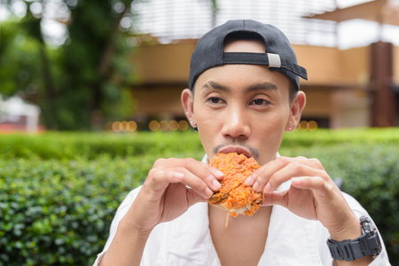 Portrait of non binary gay man eating fried chicken and drinking cola outdoors during summerの写真素材