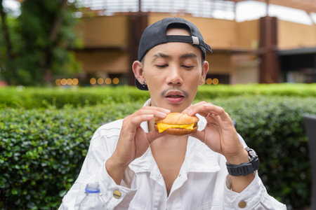 Cute Asian non binary gay man eating hamburger and drinking cola outdoors during summerの写真素材