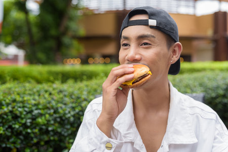 Cute Asian non binary gay man eating hamburger and drinking cola outdoors during summerの写真素材