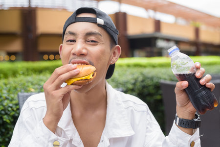 Cute Asian non binary gay man eating hamburger and drinking cola outdoors during summerの写真素材