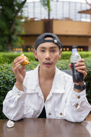 Cute Asian non binary gay man eating hamburger and drinking cola outdoors during summerの写真素材