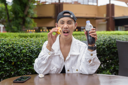 Cute Asian non binary gay man eating hamburger and drinking cola outdoors during summerの写真素材