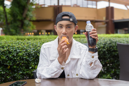 Cute Asian non binary gay man eating hamburger and drinking cola outdoors during summerの写真素材