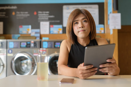 Asian transgender non-binary person sitting in laundromat laundry shop using tablet computerの写真素材
