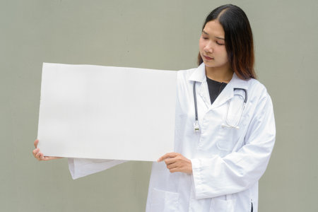 Young Asian woman doctor wearing white lab coat and stethoscope against plain backgroundの写真素材