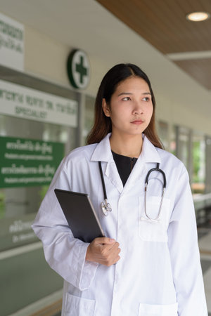 Asian young woman doctor in front of hospital clinic wearing white lab coat and stethoscopeの写真素材