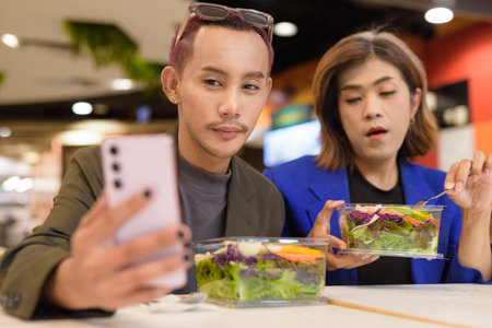 Gender diverse Asian coworker business team eating salad and working digitally taking selfie with phoneの写真素材