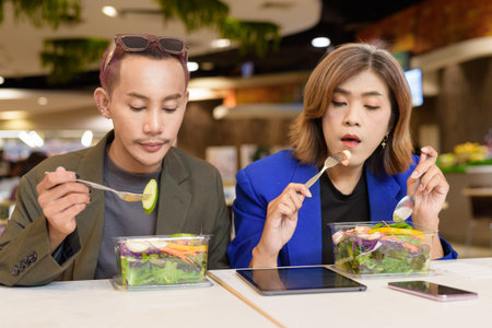 Gender diverse Asian coworkers eating salad and working digitallyの写真素材