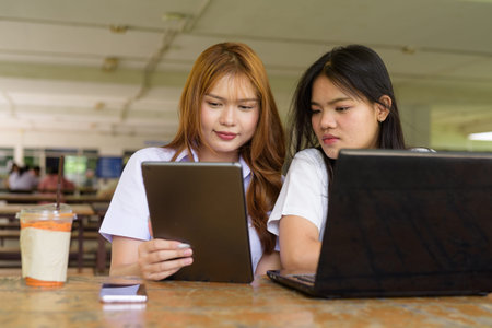 Two Thai female students sitting outdoors at a university cafe. Friendship or couple scene, casual lifestyle, education, and youth culture.の写真素材