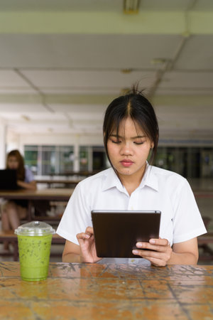 Thai teenage girl in university uniform sitting solo at a university campus coffee shop area. Relaxed lifestyle portrait showing real student life, beauty, and youth in an educational environment.の写真素材