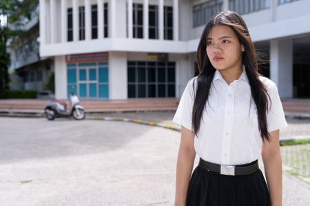 Beautiful Thai university student standing alone on campus grounds, wearing formal academic uniform. Authentic, modern education theme with focus on youth, independence, and academic ambition.の写真素材