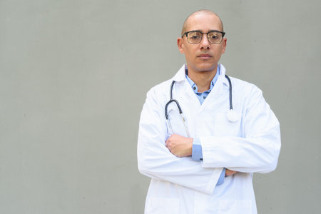 Portrait of mature bald male doctor wearing lab coat and eyeglasses with stethoscope around neck,...