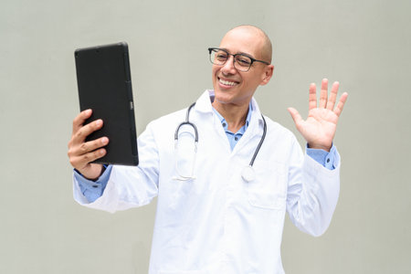 Portrait of mature bald male doctor wearing lab coat and eyeglasses with stethoscope around neck, posing against neutral wall background. Ideal for healthcare, medical, and hospital-related concepts.の写真素材