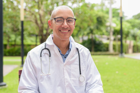 Bald male doctor in white medical uniform with stethoscope sitting outside in hospital garden. Smiling, confident, and relaxed. Concept of healthcare wellness, mental recovery, and medical lifestyle.の写真素材