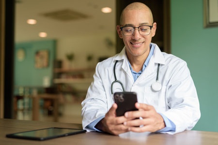 Bald male doctor in glasses and lab coat seated indoors in hospital or clinic library waiting area....