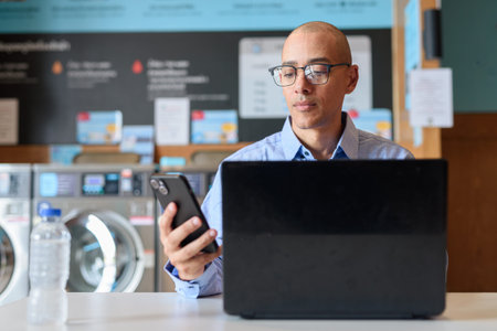 Bald male professional in blue button-down shirt working on laptop inside laundry shop. He is using...