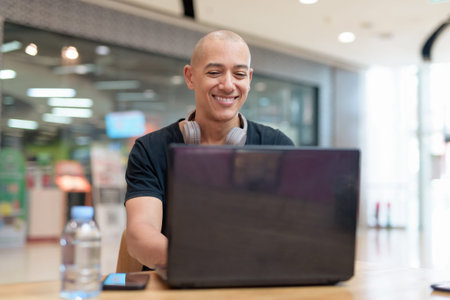 Bald adult man in black t-shirt using laptop at table inside mall food court. Water bottle on table....