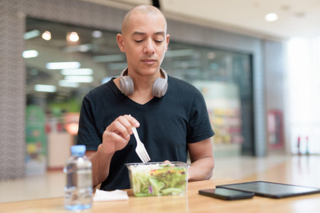 Mixed-race adult man in black t-shirt eating takeaway salad in mall food court. Healthy lifestyle,...