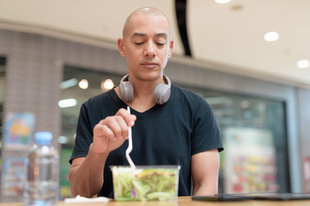Mixed-race adult man in black t-shirt eating takeaway salad in mall food court. Healthy lifestyle,...