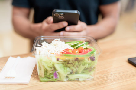 Close-up of a hand holding smartphone while interacting with fresh salad in takeaway container. Shot indoors in mall food court. Healthy eating, mobile lifestyle, and social media concept.の写真素材