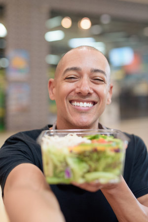 Mixed-race bald adult man in black t-shirt eating takeaway salad in mall food court. Healthy...