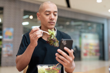 Mixed-race bald adult man in black t-shirt eating takeaway salad in mall food court. Healthy lifestyle, clean eating, and nutrition in real life setting. Natural daylight and authentic emotion.の写真素材