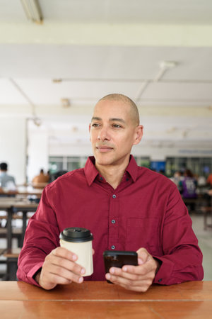 Stylish adult man in burgundy button-down shirt using mobile phone while sitting alone at table in outdoor public area. Modern digital lifestyle concept with takeaway coffee cup.の写真素材