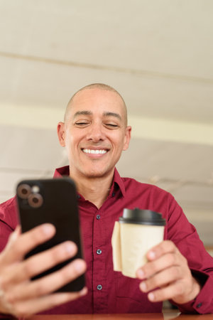 Stylish adult man in burgundy button-down shirt using mobile phone while sitting alone at table in outdoor public area. Modern digital lifestyle concept with takeaway coffee cup.の写真素材