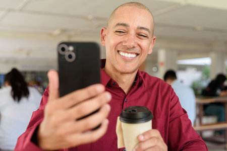 Stylish adult man in burgundy button-down shirt using mobile phone while sitting alone at table in outdoor public area. Modern digital lifestyle concept with takeaway coffee cup.の写真素材