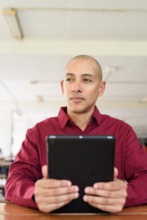 Confident man wearing burgundy colored shirt using tablet computer at coffee shop table outdoors in...
