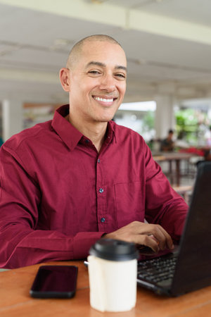 Bald adult man in burgundy button-down shirt working on laptop while sitting at outdoor coffee shop...