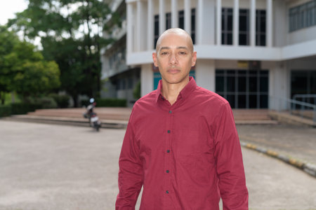 Stylish bald adult Italian man in burgundy button-down shirt posing confidently in front of modern university building during sunny summer day. Urban academic lifestyle, education success concept.の写真素材