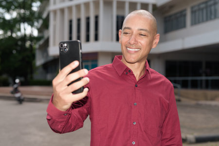 Bald Italian man in burgundy shirt checking his smartphone while standing in front of university...