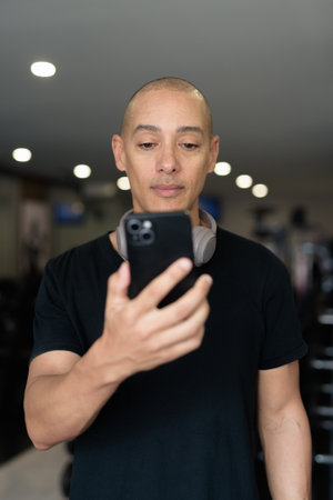 Bald Hispanic man training in gym for fitness and workout. Muscular male exercising indoors with confidence, wearing black shirt and headphones. Healthy lifestyle, strength and wellness focus.の写真素材