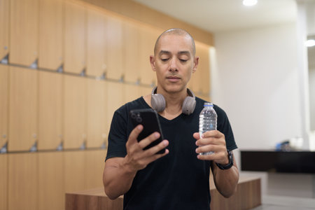 Latin adult male in locker room holding water bottle while using smartphone. Gym workout recovery, hydration habit, digital fitness routine, privacy and wellness lifestyle after training.の写真素材