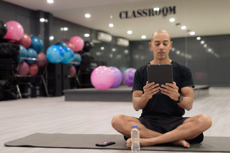 Bald Hispanic man training in gym for fitness and workout. Muscular male using digital tablet computer indoors with confidence, wearing black shirt. Healthy lifestyle, strength and wellness focus.の写真素材