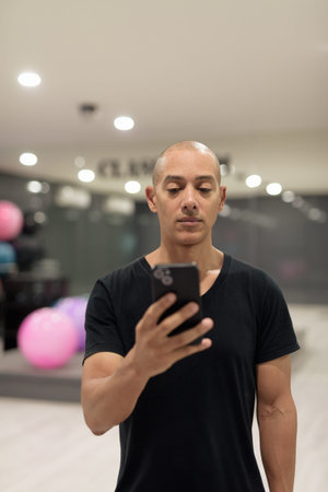 Hispanic man using mobile phone in gym for fitness and workout. Muscular male exercising indoors with confidence, wearing black shirt. Healthy lifestyle, strength and wellness focus.の写真素材