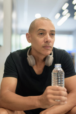Hispanic bald man athlete in gym sitting after workout relaxing and holding water bottle ready to...