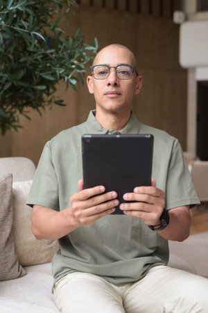 Stylish bald Hispanic man browsing tablet in elegant indoor cafe, hotel bar or restaurant. Digital...