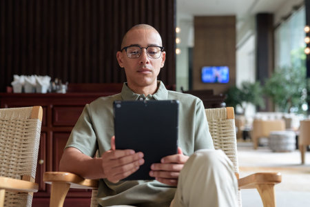 Latin adult man in eyeglasses using tablet while seated in hotel lobby or bar lounge. Modern digital...