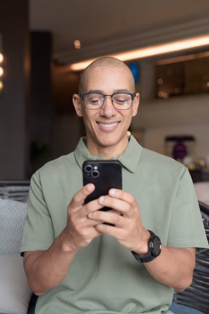 Latin adult man in eyeglasses using phone while seated in hotel lobby or bar lounge. Modern digital...