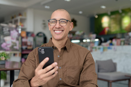 Bald Latin man in eyeglasses sitting at cafe using smartphone. Wearing casual shirt, smiling and confident. Modern digital lifestyle, communication, mobile technology, urban coffee shop scene.の写真素材