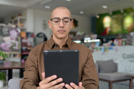 Smiling bald man in his 40s using tablet device while sitting in cozy cafe. Wearing eyeglasses and casual button shirt. Technology, digital habits, and leisure lifestyle theme.の写真素材