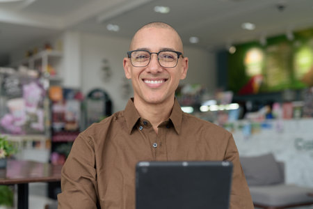 Smiling bald man in his 40s using tablet device while sitting in cozy cafe. Wearing eyeglasses and...