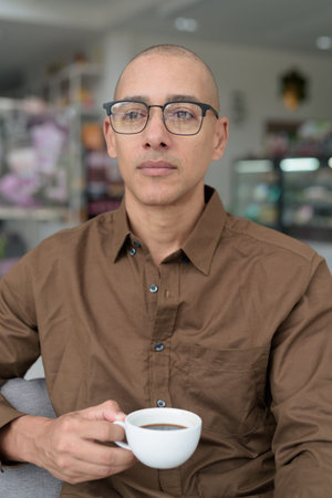Bald hispanic man in his 40s sitting in coffee shop, wearing casual shirt and eyeglasses. Confident and happy expression. Urban lifestyle, modern man, adult portrait in relaxed environment.の写真素材