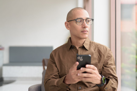 Bald Latin man in eyeglasses sitting at cafe using smartphone. Wearing casual shirt, smiling and confident. Modern digital lifestyle, communication, mobile technology, urban coffee shop scene.の写真素材